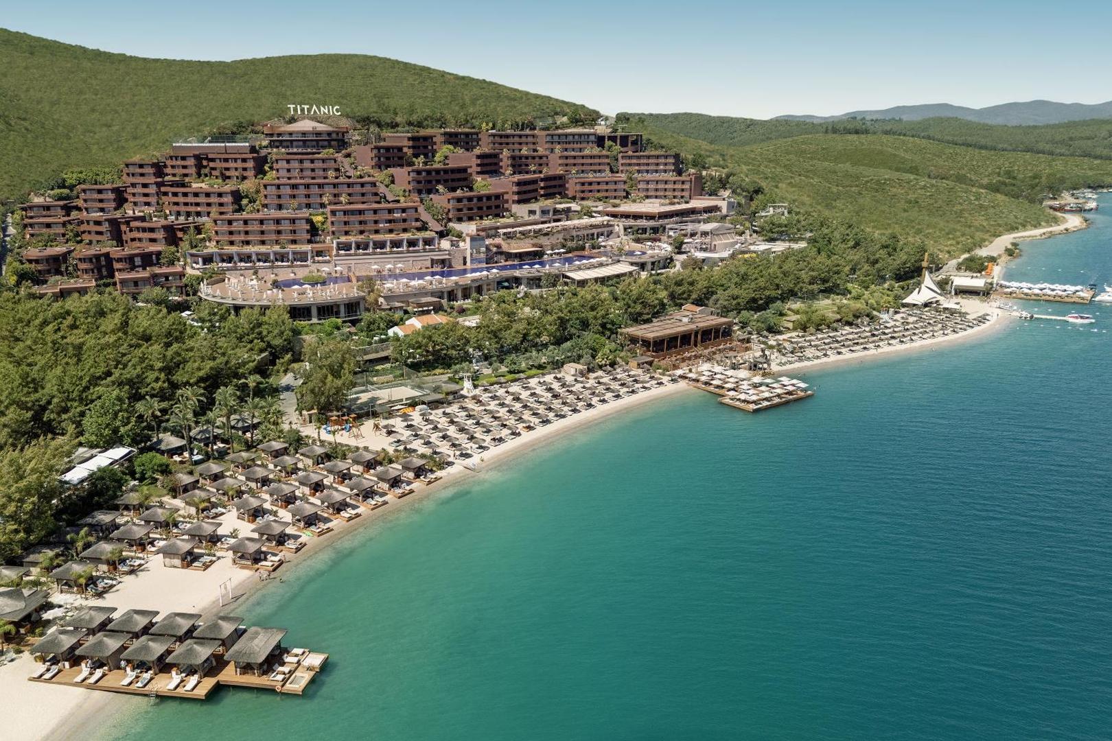 Panorāmas skats uz Titanic Luxury Collection Bodrum viesnīcu kalna nogāzē pie tirkīzzila līča ar privāto pludmali un piestātnēm