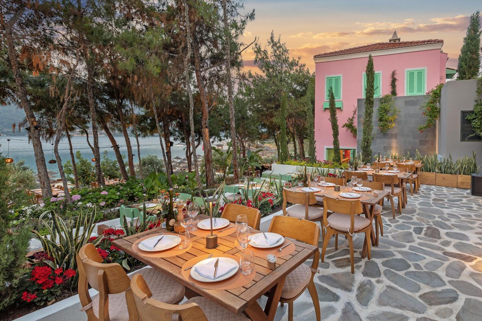 Viesnīcas Titanic Luxury Collection Bodrum āra restorāns ar koka galdiem, ziediem un rozā ēku fonā saulrieta laikā piekrastē