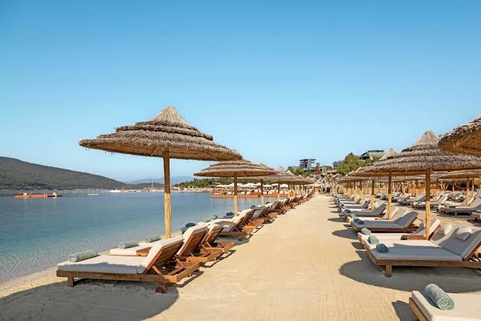 Titanic Luxury Collection Bodrum pludmales zona ar koka zviļņiem un salmu saulessargiem uz gaišām smiltīm pie mierīgas jūras