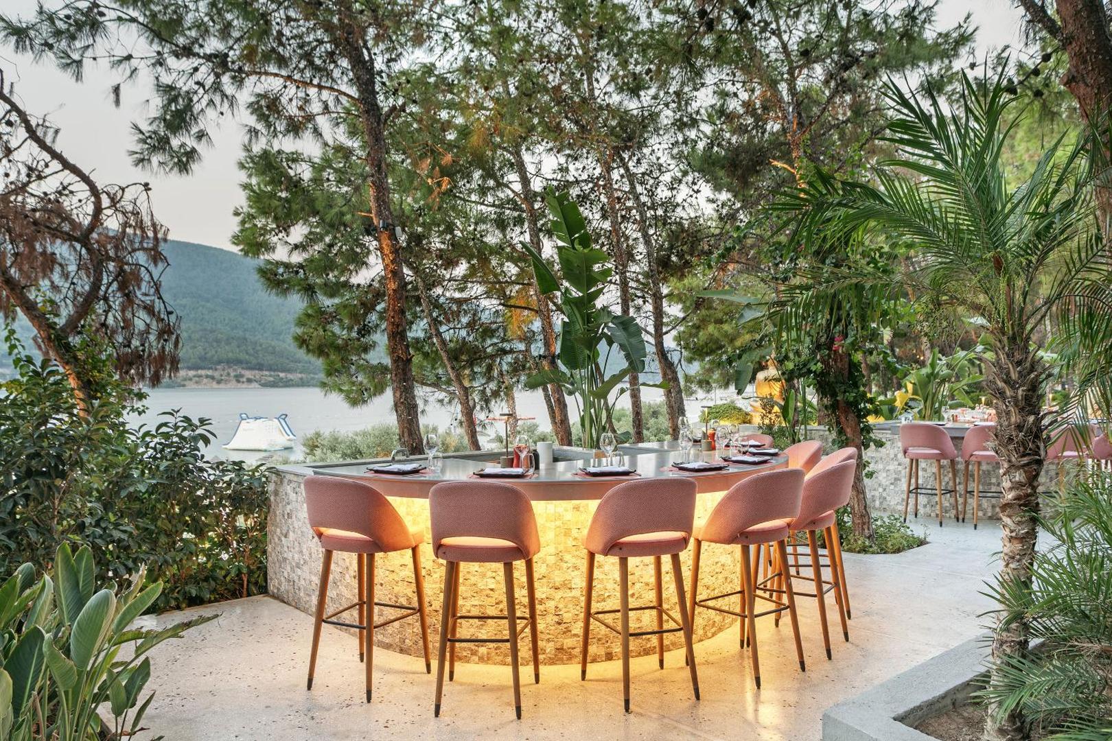 Titanic Luxury Collection Bodrum āra restorāna bāra zona ar rozā krēsliem, apgaismojumu un skatu uz jūru caur priedēm