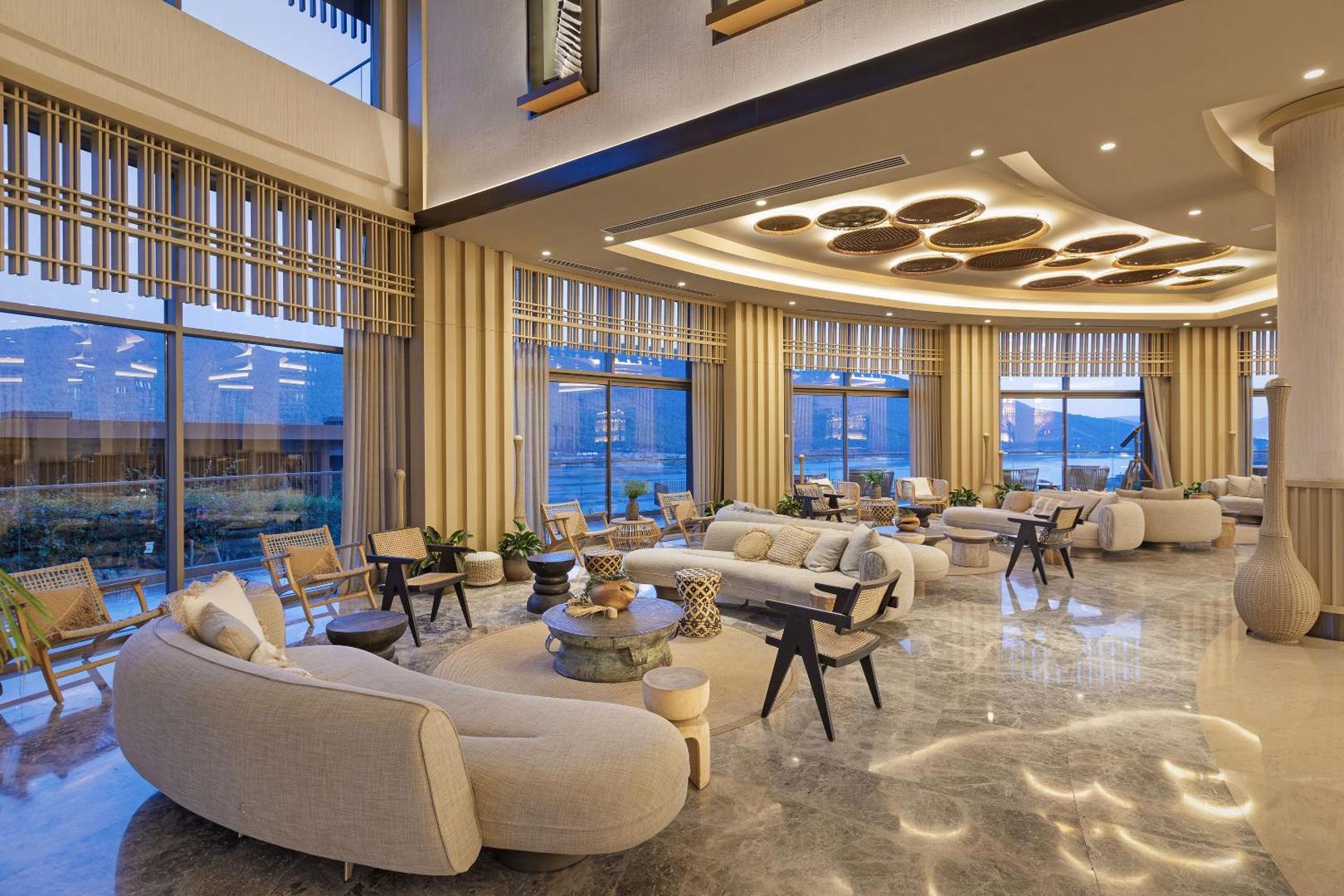 Stilīga atpūtas zona Titanic Luxury Collection Bodrum lobby ar mīkstajiem dīvāniem un lieliem logiem uz piekrasti