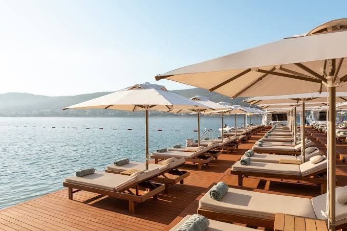 Titanic Luxury Collection Bodrum koka sauļošanās piestātne ar elegantiem saulessargiem un ērtiem zviļņiem pie dzidrajiem Egejas jūras ūdeņiem