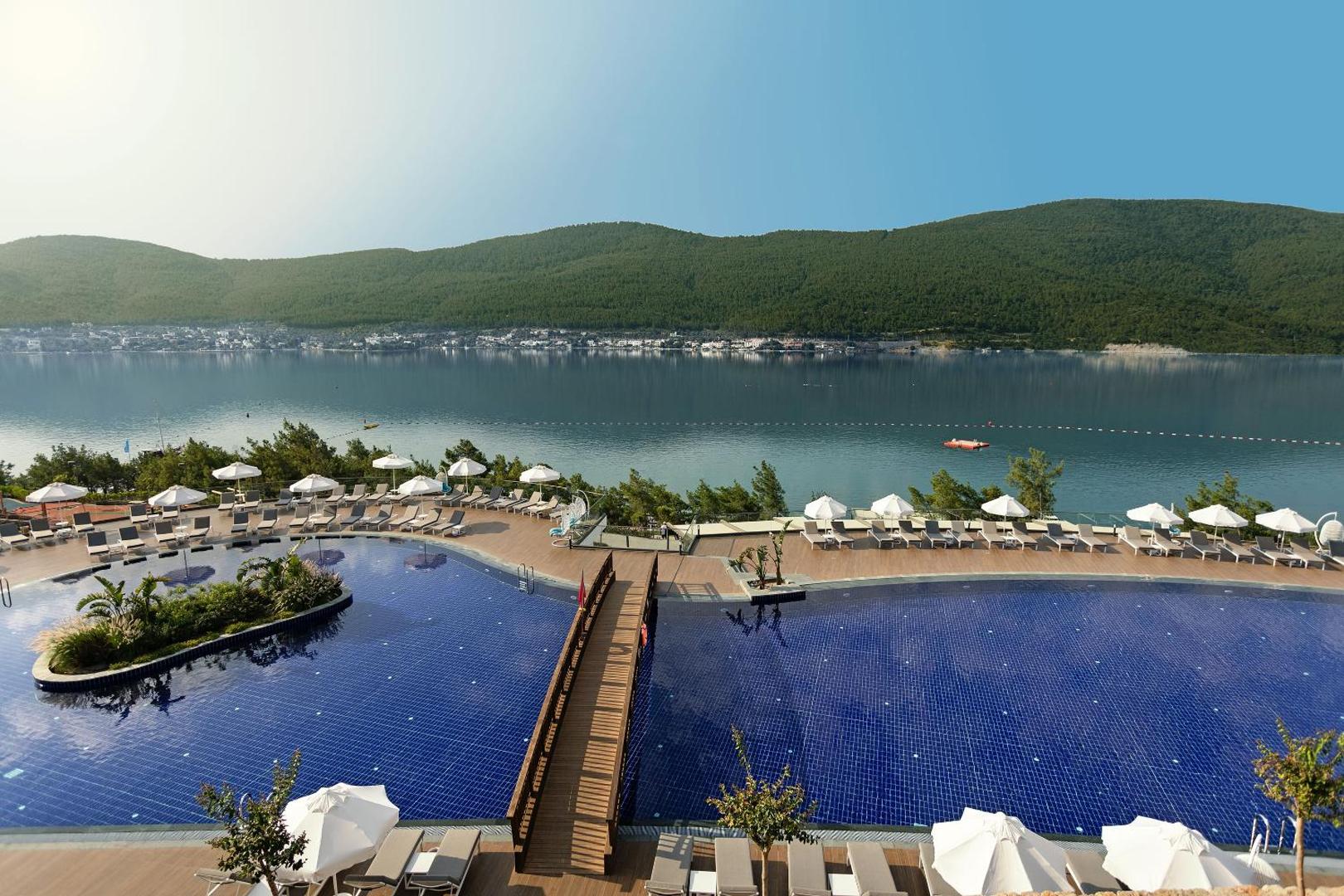 Titanic Luxury Collection Bodrum galvenais āra baseins ar koka tiltiņu, saulessargiem un skatu uz Egejas jūras līci un kalniem