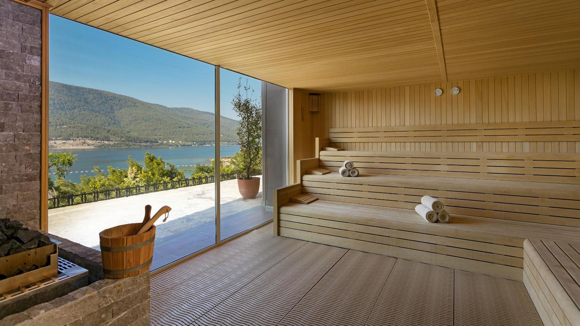 Moderna sauna ar panorāmas logu Titanic Luxury Collection Bodrum viesnīcā, no kuras paveras skats uz jūru un kalniem