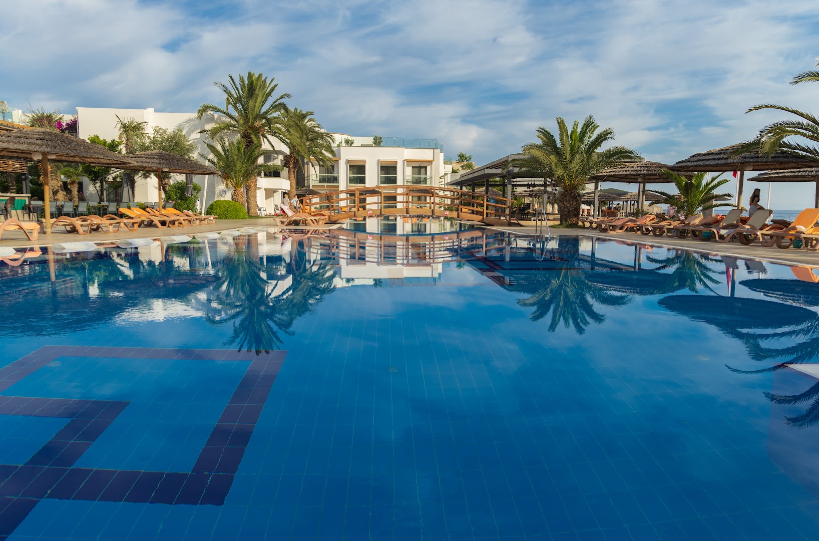 Samara Hotel Bodrum galvenais baseins ar koka tiltiņu, saulessargiem un palmām gaišā dienas laikā Turcijā