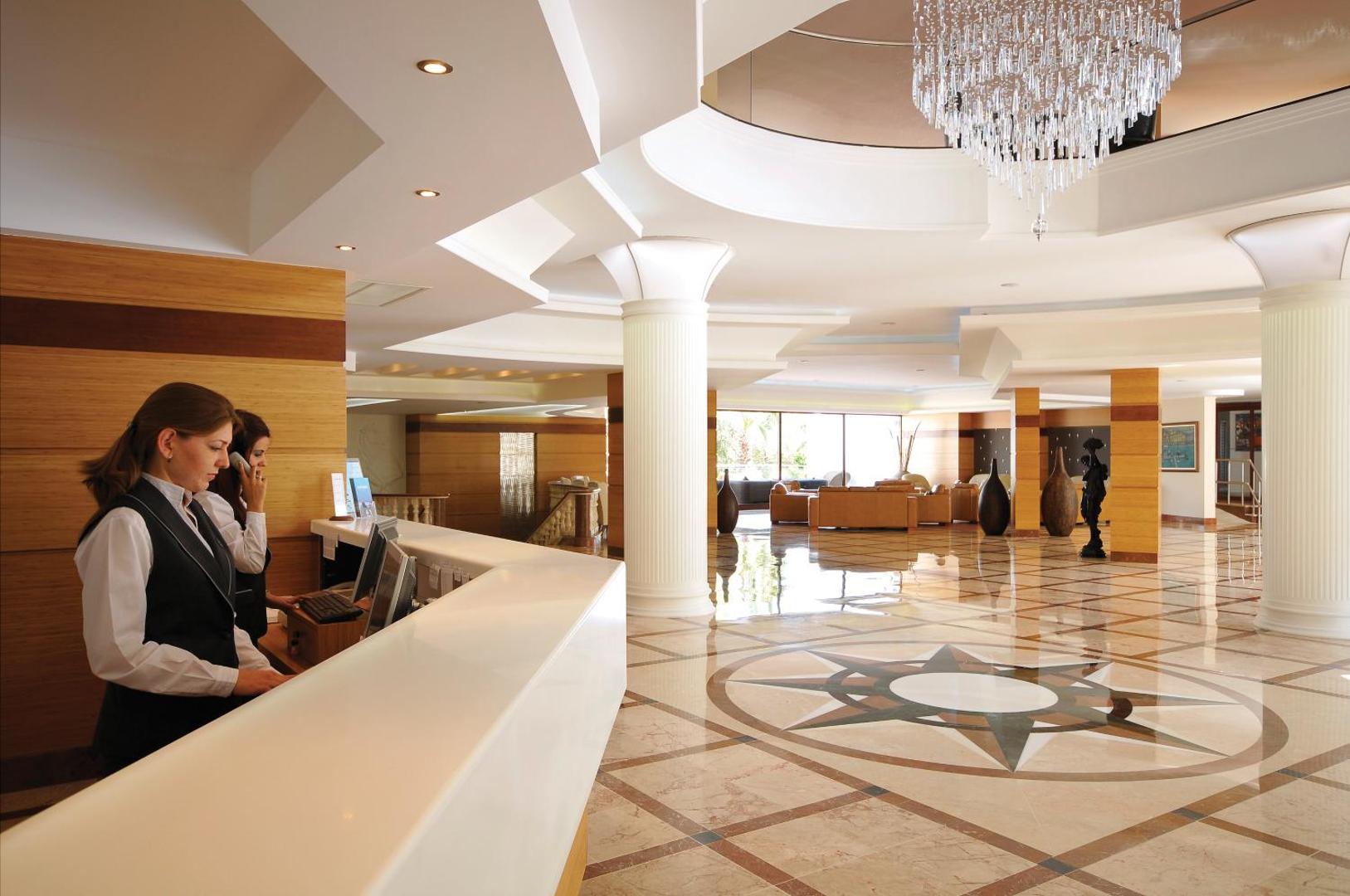 Samara Hotel Bodrum plašais vestibils ar marmora grīdu, kristāla lustru un recepcijas darbiniekiem, kuri apkalpo viesus pie baltas letes.