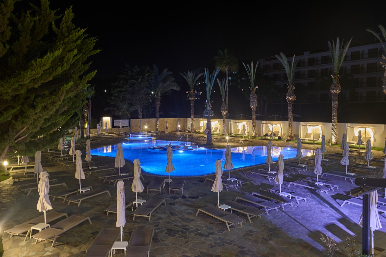 Bohemian Gardens Hotel baseina zona vakarā — izgaismots zils baseins, daudzi sauļošanās krēsli un palmas nakts apgaismojumā.