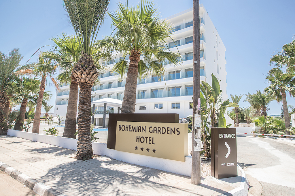 Bohemian Gardens Hotel 4* viesnīcas izkārtne pie iebrauktuves, fonā galvenā ēka un augstas palmas skaidrā dienas laikā