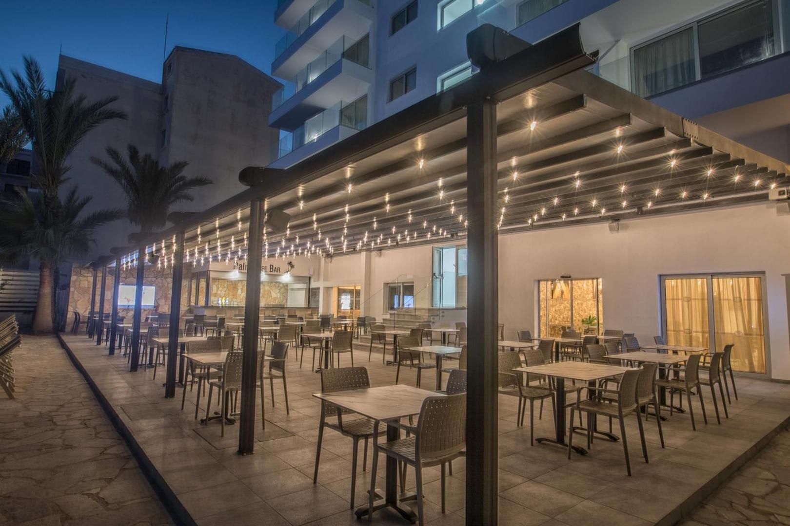 Bohemian Gardens Hotel bāra Palm Tree Bar āra terase vakarā — galdiņi zem nojumes ar dekoratīvām lampiņu virtenēm.