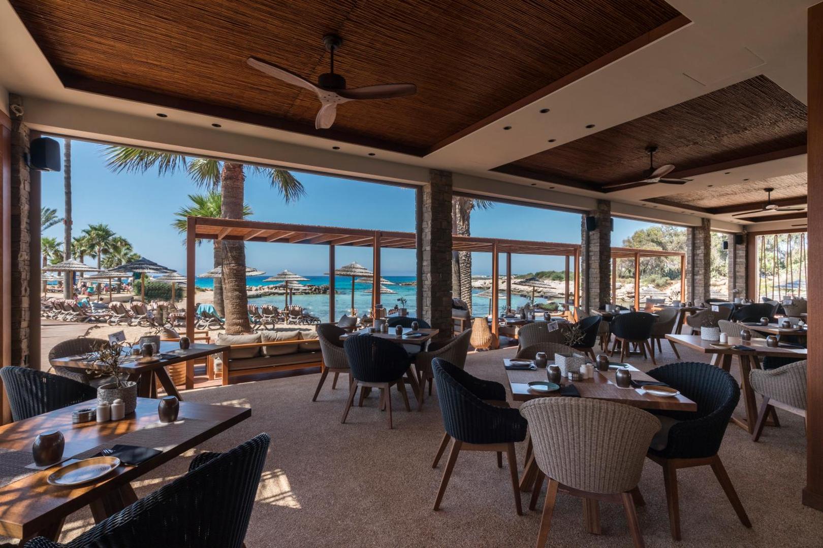 Adams Beach viesnīcas restorāna terase ar skatu uz pludmali — koka griesti, eleganti galdi un palmas fonā