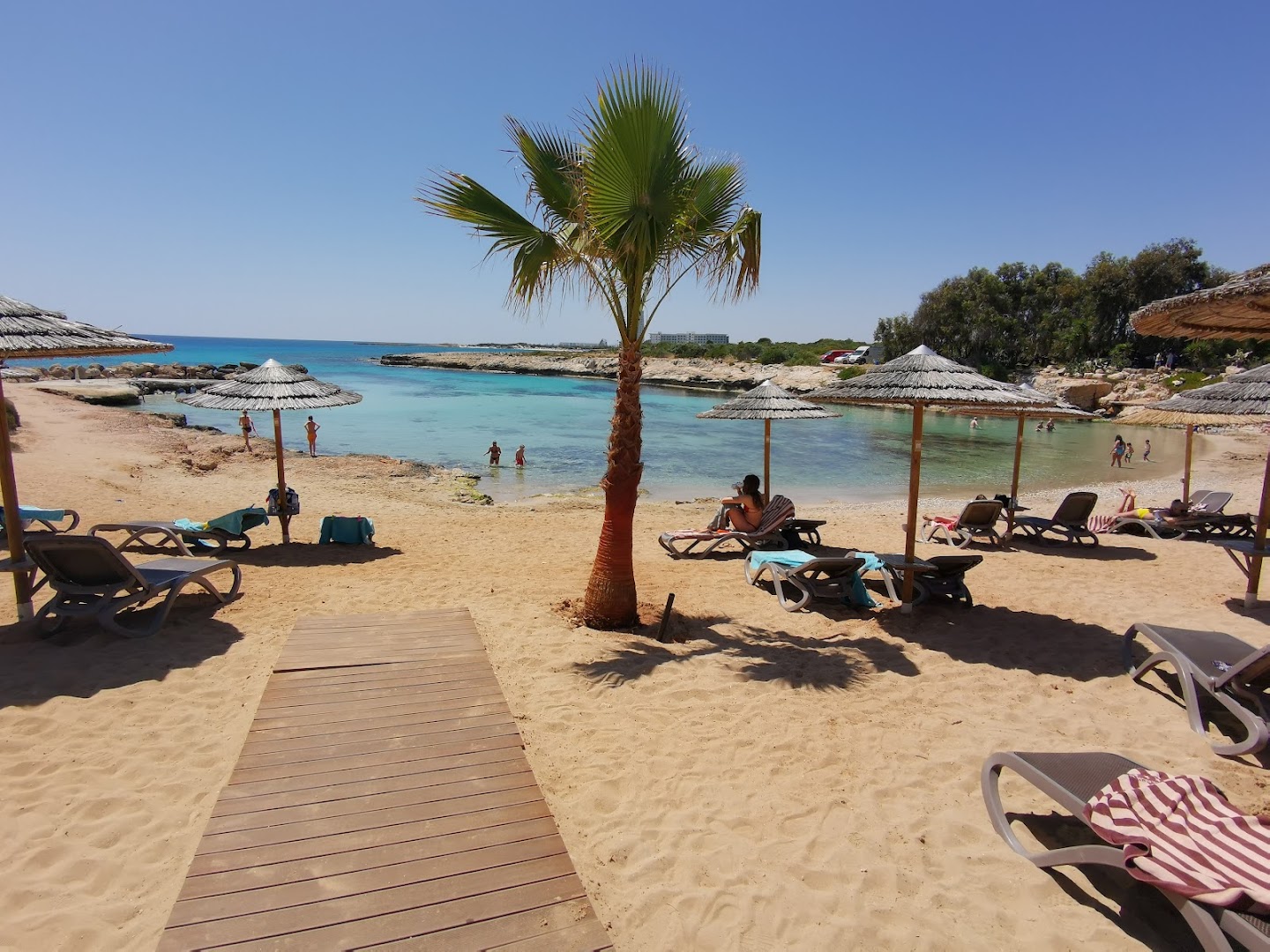 Adams Beach viesnīcas privātā smilšu pludmale Aijanapā, Kiprā — tirkīzzila līča ūdeņi, saulessargi un atpūtas krēsli mierīgai atpūtai pie jūras