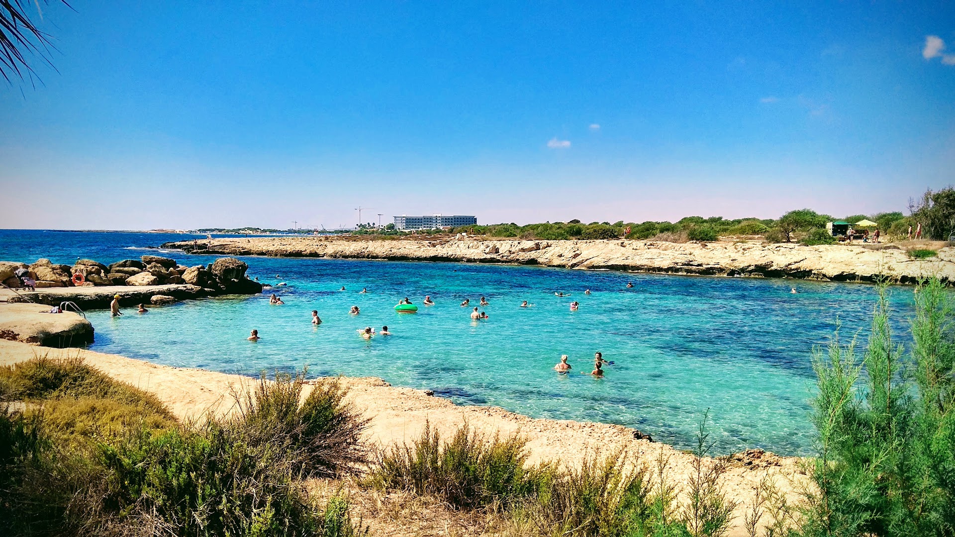 Adams Beach viesnīcas privātā lagūna Aijanapā, Kiprā — tirkīzzils ūdens, smilšu pludmale un atpūtnieki peldoties saulainā dienā