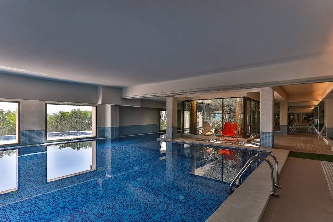 Carine Hotel Park iekštelpu baseins ar panorāmas logiem un skatu uz dārzu — moderna spa zona ar atpūtas krēsliem