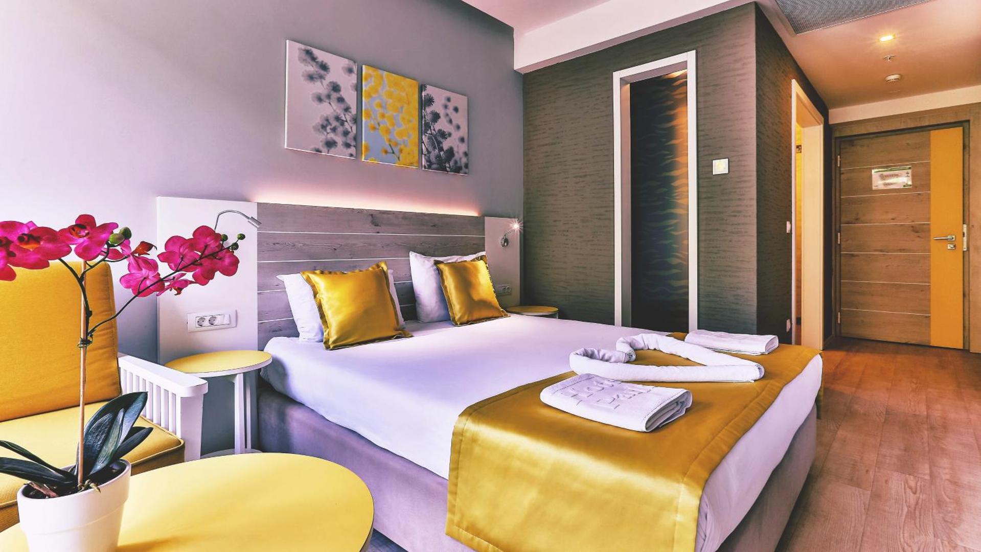 Carine Hotel Park viesnīcas numurs ar divguļamo gultu, dzelteniem dekoratīviem elementiem un ziedu kompozīciju priekšplānā