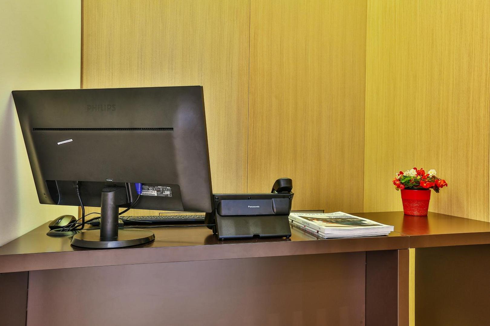 Viesnīcas Bono darba zona ar datoru, telefonu un žurnāliem uz tumša koka galda, piemērota biznesa ceļotājiem.
