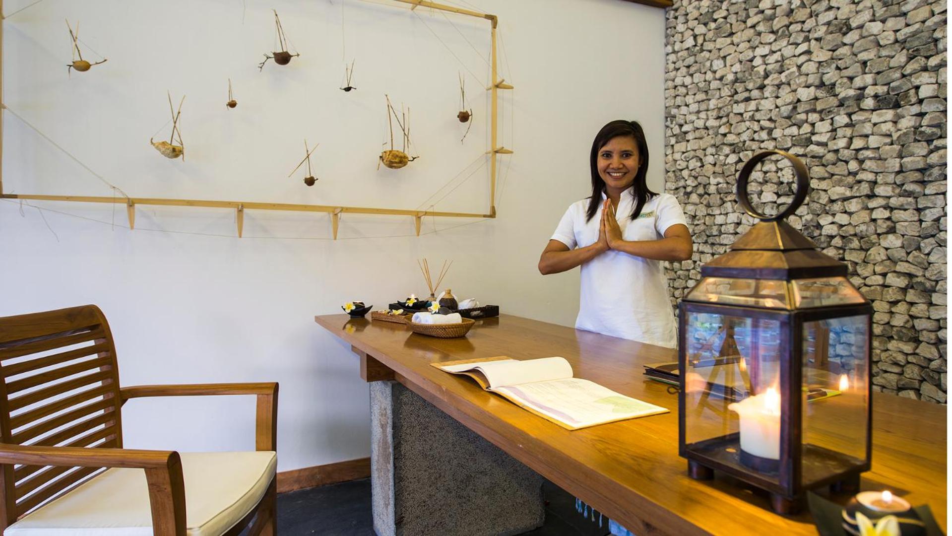 The Barefoot Eco Hotel viesmīlīga darbiniece pie koka recepcijas galda Maldīvu salās — gaišs interjers ar akmens sienu