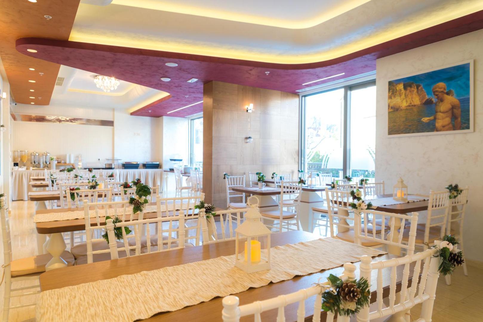 Hotel Kadmo restorāna zāle ar balti klātiem galdiem, dekoratīviem elementiem un bufetes zonu brokastīm