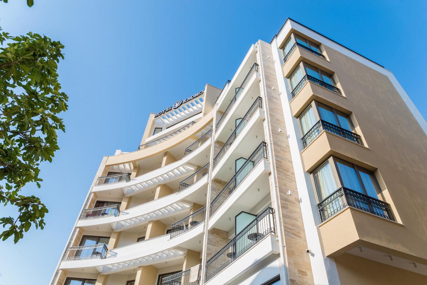 Hotel Kadmo by Aycon modernā fasāde ar gaišiem balkoniem un stiklotiem logiem pret zilu debesu fonu Budvā