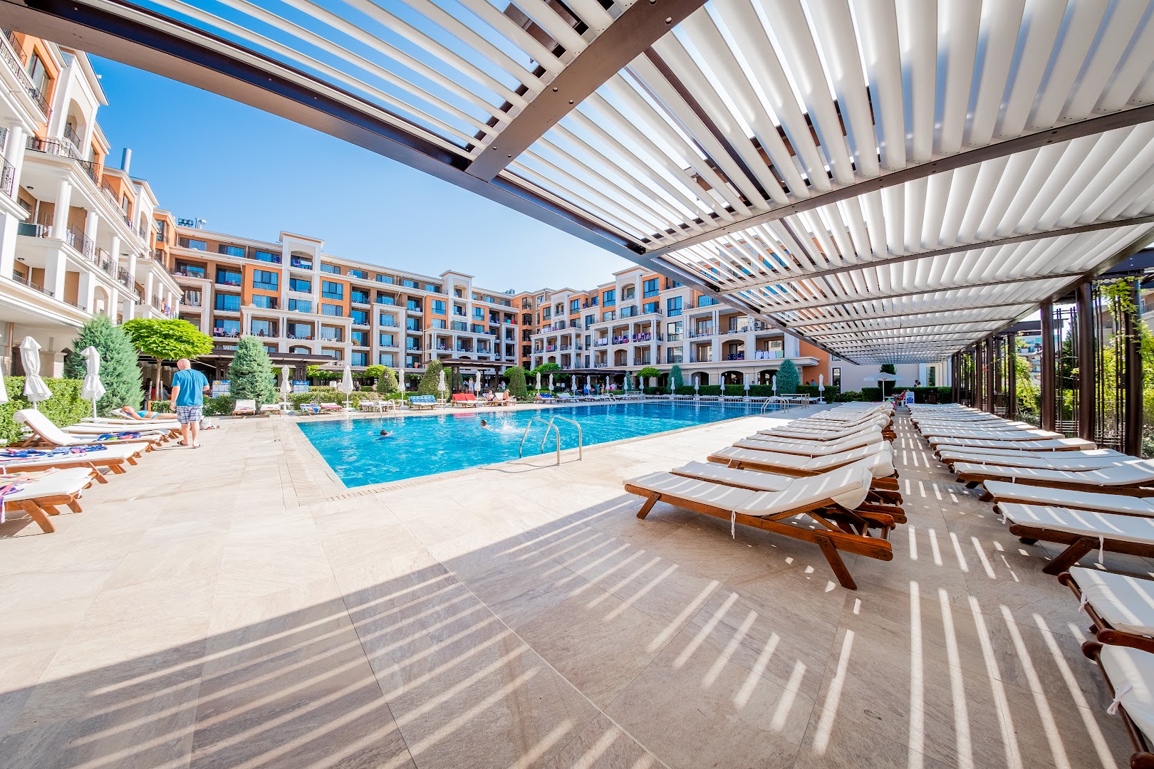 Atpūtas zona pie Premier Fort Beach Hotel baseina ar koka sauļošanās krēsliem zem nojumes un skatu uz viesnīcas ēku