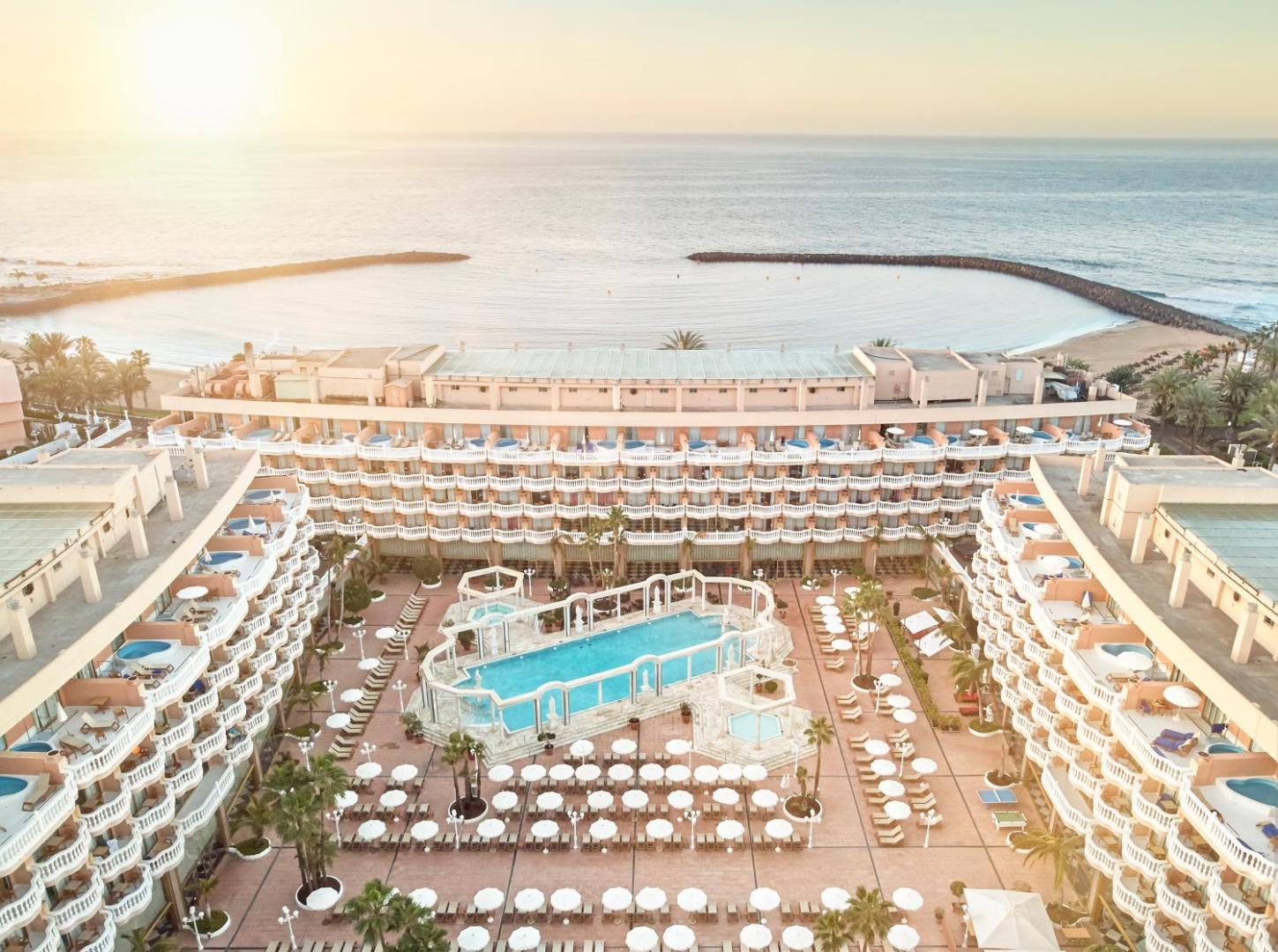 Cleopatra Palace viesnīcas panorāmas skats Tenerifē ar plašu āra baseinu, saulessargiem un skatu uz okeāna līci saulrietā