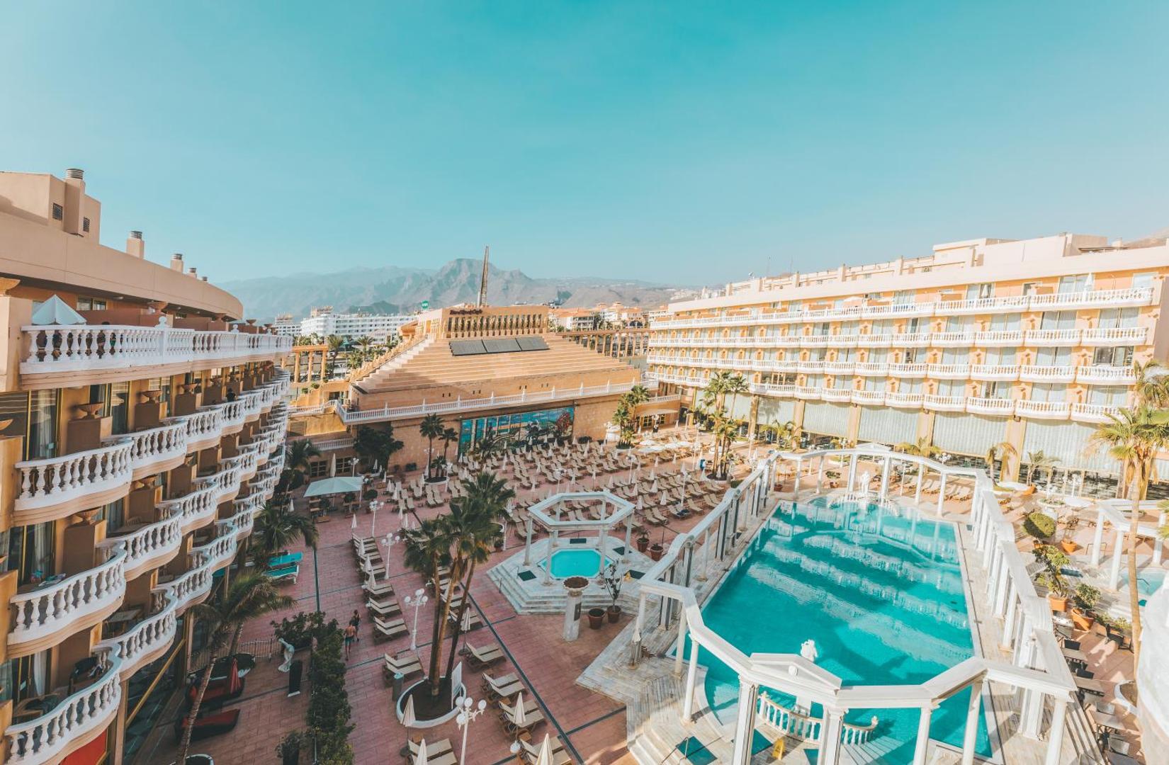 Cleopatra Palace viesnīcas centrālais baseins ar kolonnām, sauļošanās krēsliem un skatu uz kalniem Tenerifes kūrortā.