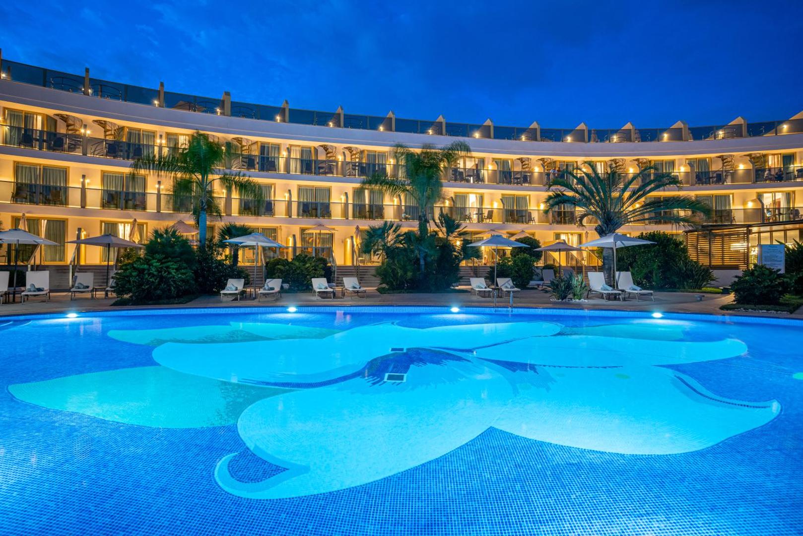 Sir Anthony Hotel izgaismots āra baseins vakarā Tenerifē ar skatu uz viesnīcas fasādi, balkoniem un palmām piekrastē