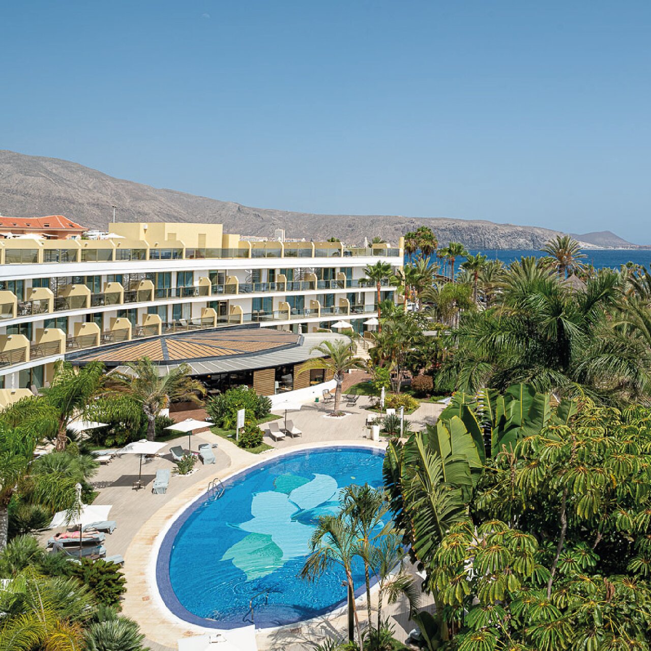 Sir Anthony Hotel teritorijas skats no augšas ar baseinu, palmām un kalniem fonā pie okeāna Tenerifes dienvidos