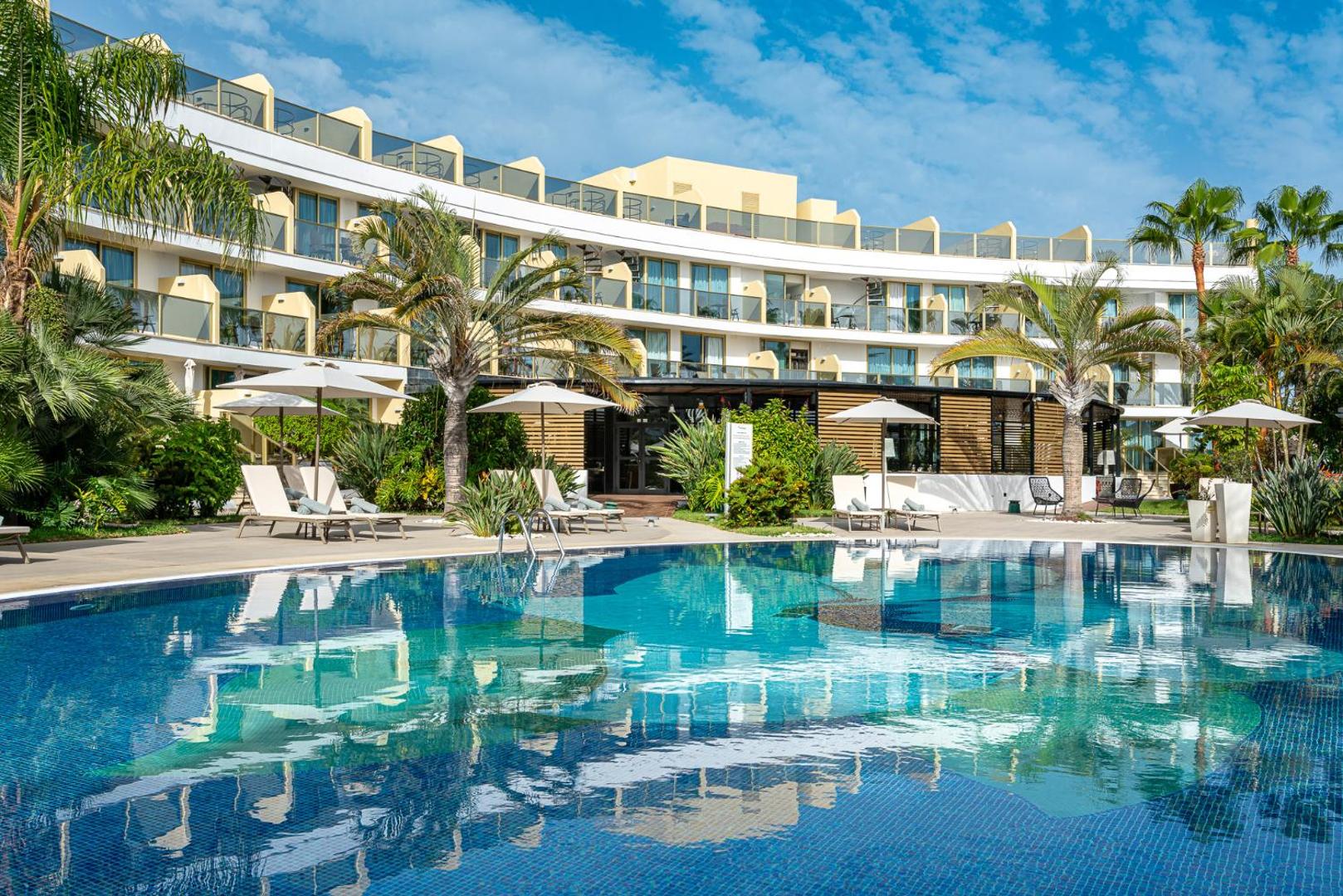 Sir Anthony Hotel ēkas fasāde un lielais āra baseins Tenerifē, saulaina diena ar palmām un atpūtas krēsliem