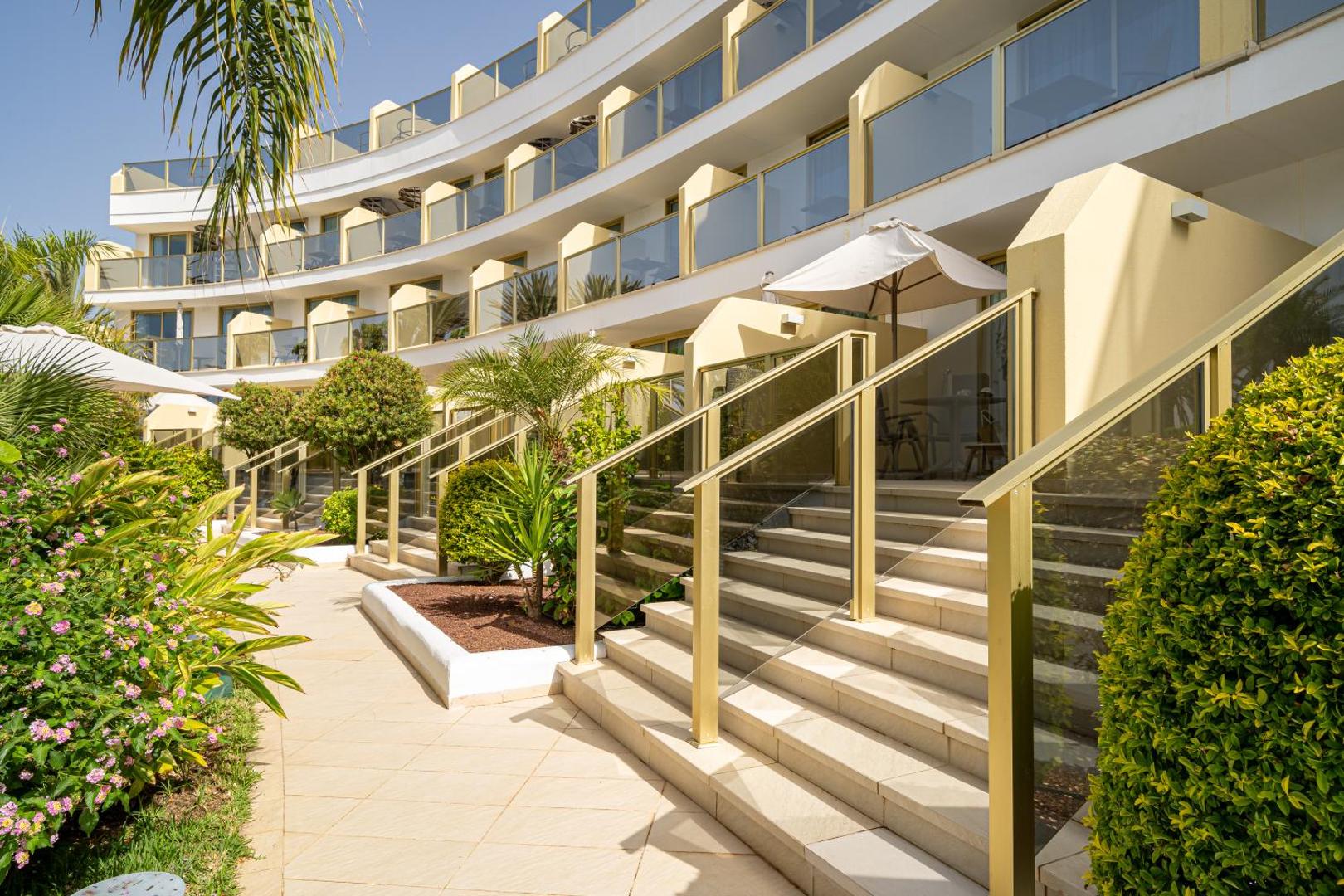 Sir Anthony Hotel modernā fasāde ar pusapaļiem balkoniem, kāpnēm un koptu dārzu ar eksotiskiem augiem un palmām