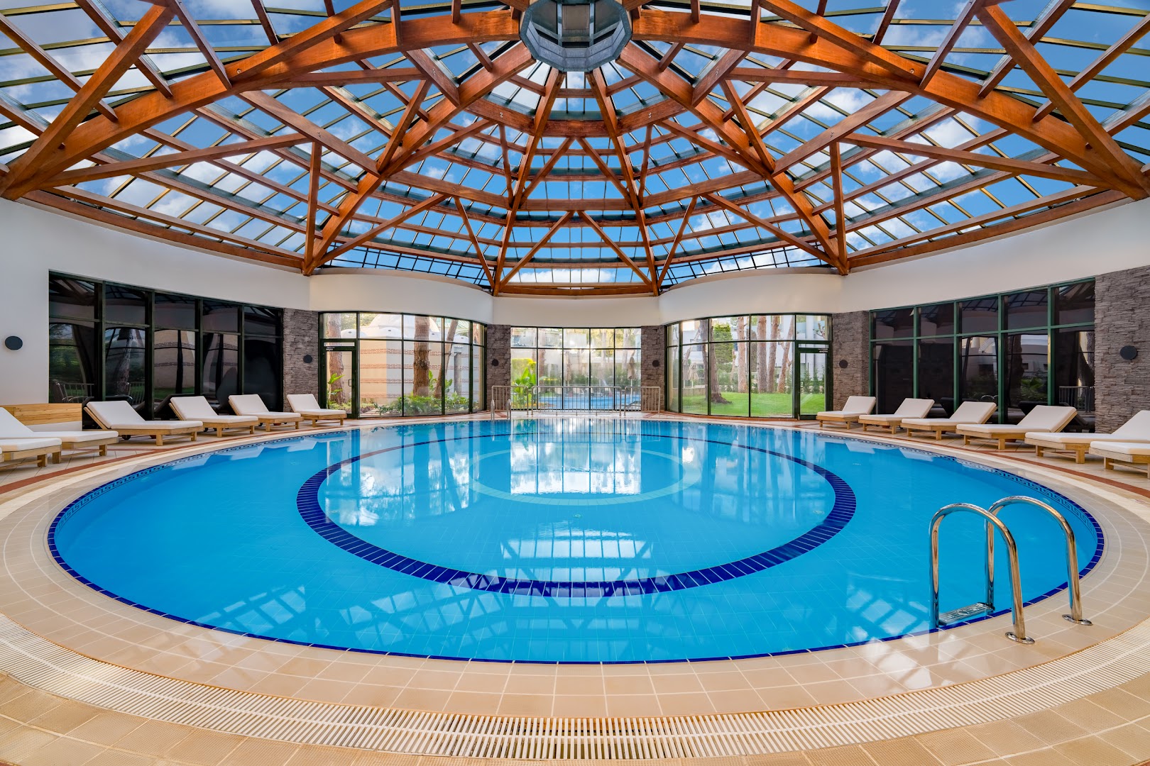 Rixos Park Belek apaļš iekštelpu baseins zem iespaidīga koka un stikla kupola ar atpūtas krēsliem spa zonā.