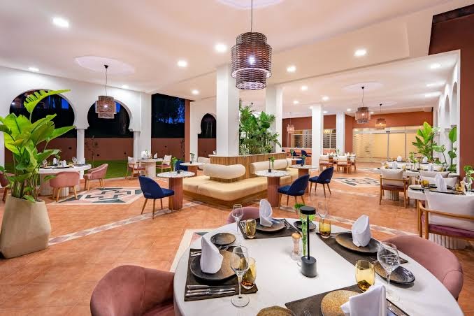 Rixos Park Belek iekštelpu restorāns ar modernu dizainu, rozā krēsliem, dekoratīviem augiem un stilīgiem gaismekļiem