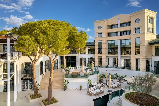 Rixos Park Belek viesnīcas pagalms ar atpūtas zonu, galdiņiem un lielām priedēm saulainā dienā Belekas reģionā, Turcijā