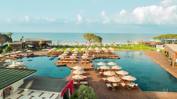 Rixos Park Belek panorāmas skats uz baseinu kompleksu un Vidusjūras piekrasti saulainā dienā Belekas kūrortā.