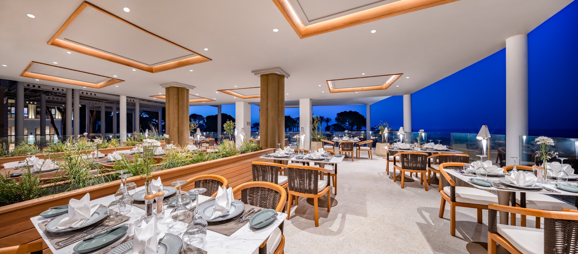 Rixos Park Belek elegants restorāns ar klātiem galdiem, pītiem krēsliem un atvērtu skatu uz vakara debesīm