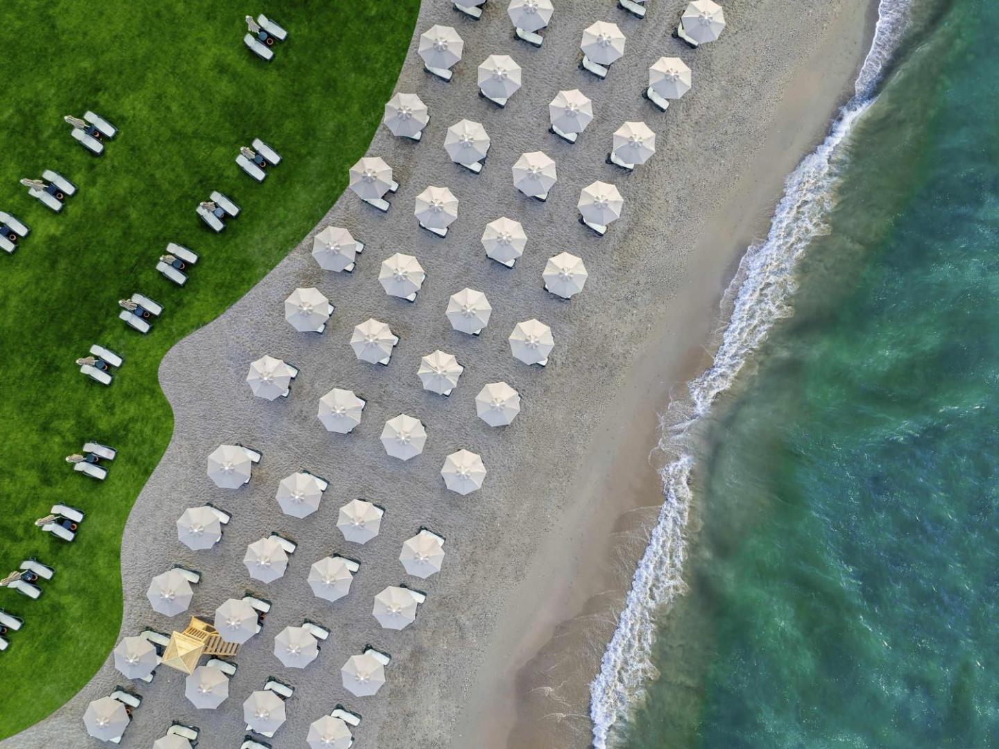 Rixos Park Belek privātā pludmale no putna lidojuma — simetriskas saulessargu rindas smiltīs blakus zaļam zālienam un tirkīzzilai jūrai