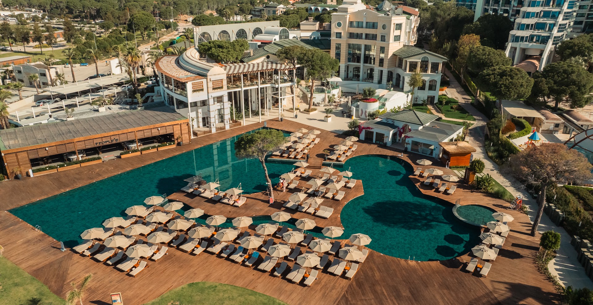 Rixos Park Belek viesnīcas ēkas un lielais āra baseins ar daudziem saulessargiem un koka klāju Belekā, Turcijā.