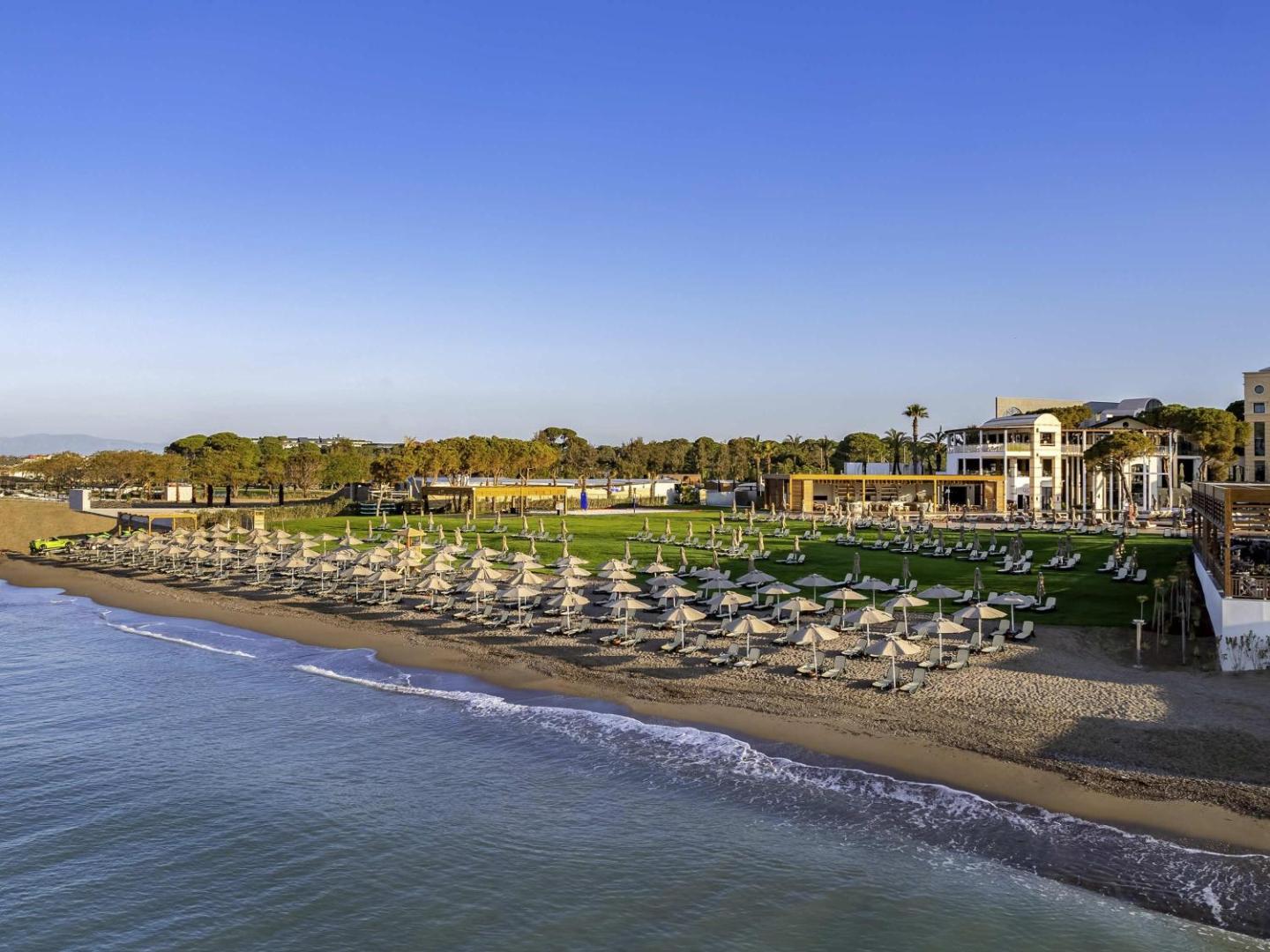 Rixos Park Belek viesnīcas ēkas un plašā pludmales zona saulrietā — skats uz piekrasti, zālienu un atpūtas krēsliem Belekā
