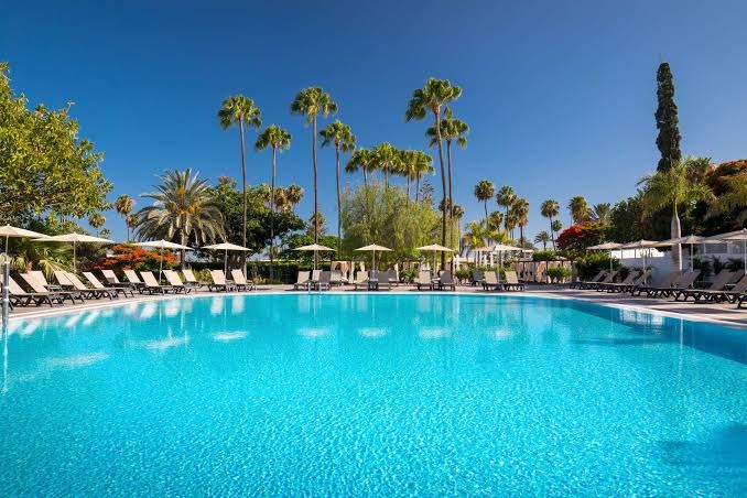 Barcelo Margaritas baseins ar rindu saulessargiem un augstām palmām pret zilām debesīm, radot tropisku atmosfēru Grankanārijas viesnīcā.