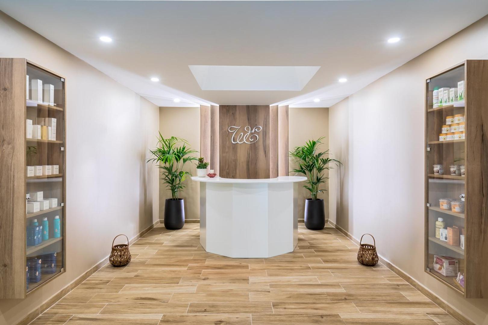 Mūsdienīga Barcelo Margaritas spa recepcijas zona ar gaišu koka apdari, dekoratīviem augiem un produktu vitrīnām