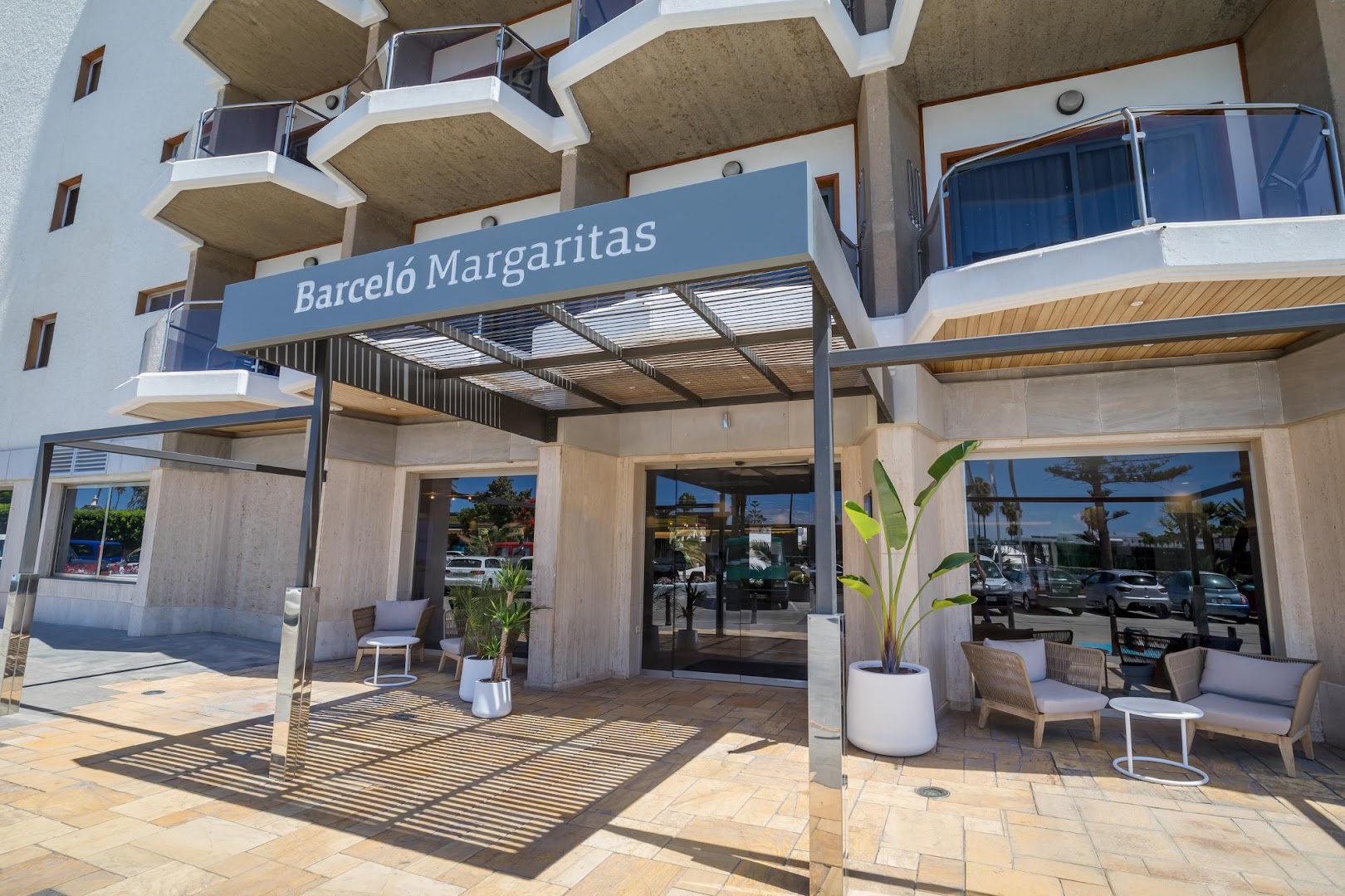 Barceló Margaritas galvenā ieeja ar viesnīcas nosaukumu uz nojumes, āra mēbelēm un modernu stikla fasādi