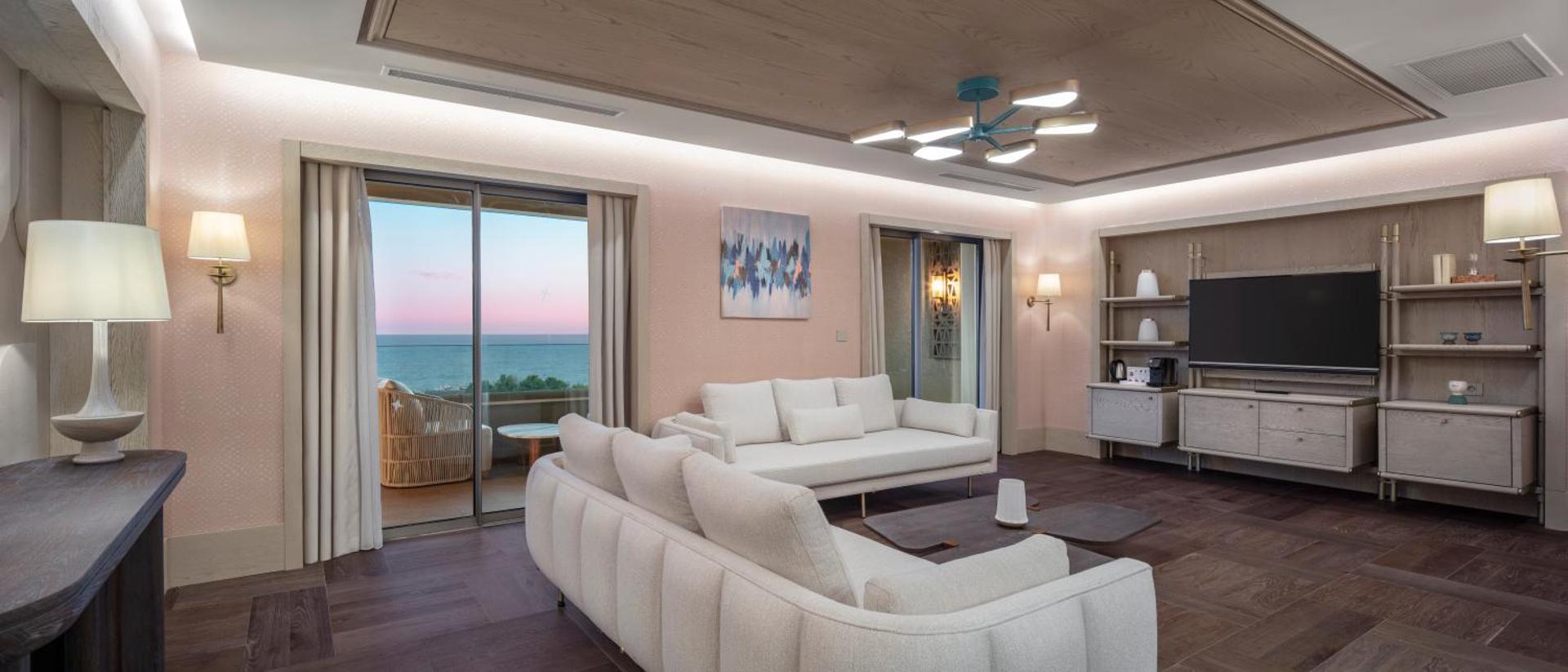 Ela Excellence Resort Belek viesistabas numurs ar gaišiem dīvāniem, koka griestiem un skatu uz jūru saulrietā Belekā, Turcijā.