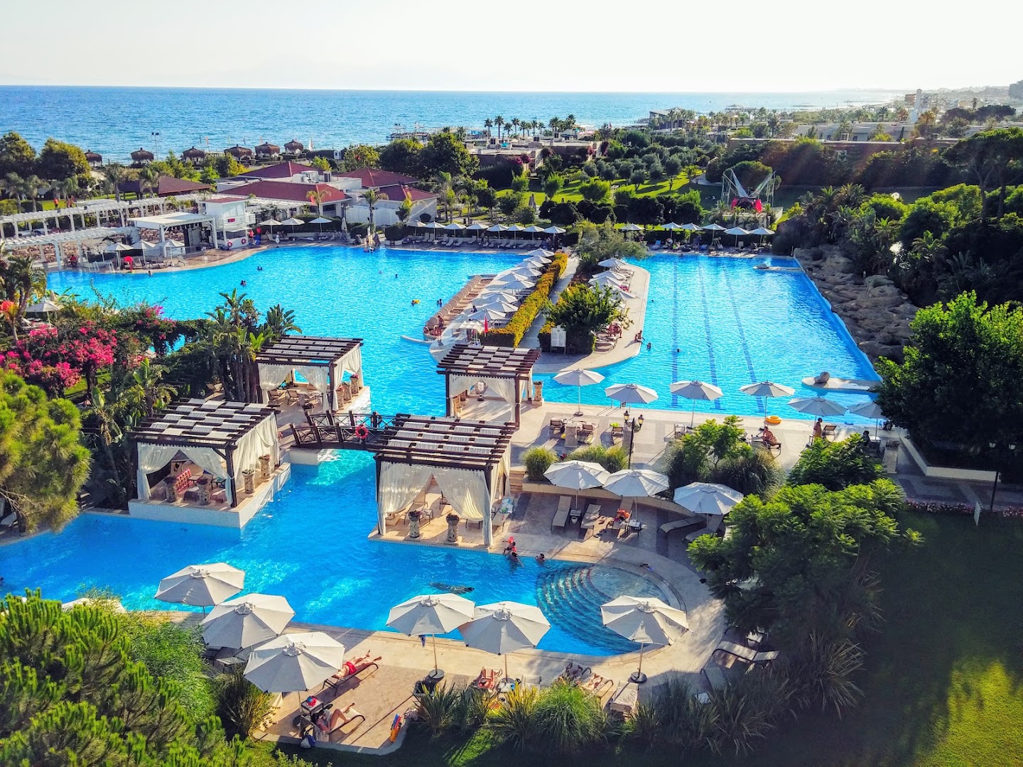Ela Excellence Resort Belek galvenais baseins ar greznām atpūtas nojumēm, tiltiņiem un skatu uz Vidusjūru saulrietā