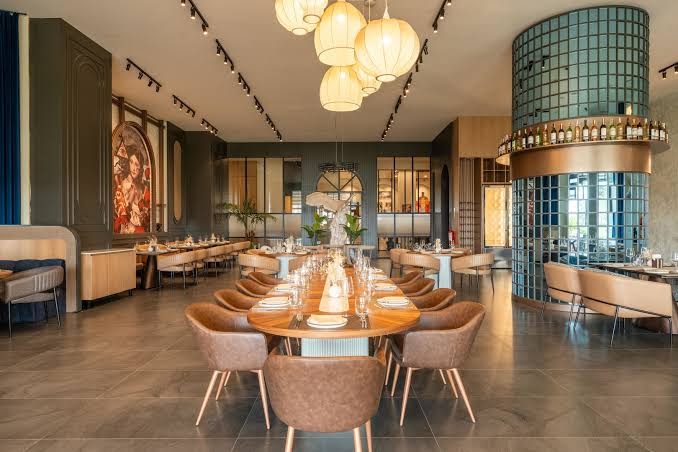 Ethno Belek restorāna interjers ar modernām lustrām, gariem koka galdiem, ādas krēsliem un dekoratīvu stikla kolonnu centrā