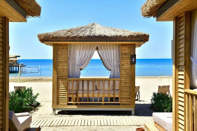 Ethno Belek viesnīcas koka bungalo pludmalē ar salmu jumtu un baltiem aizkariem, skats uz zilo Vidusjūru un smilšu krastu Turcijā