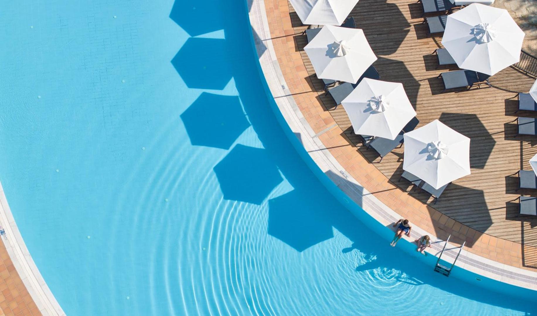 Agapi Beach Resort galvenais baseins no putna lidojuma — zils ūdens, balti saulessargi un koka terase ar atpūtas krēsliem Krētā