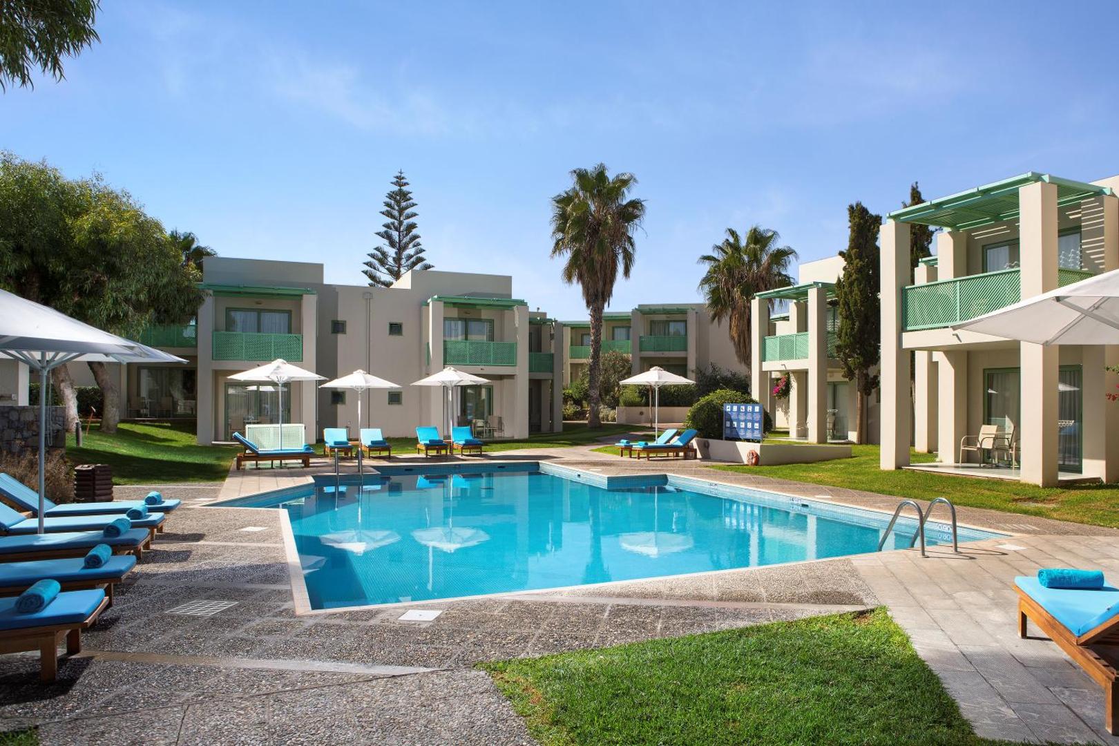 Agapi Beach Resort bungalo tipa numuri pie neliela baseina — zāliens, palmas un atpūtas krēsli ar ziliem dvieļiem