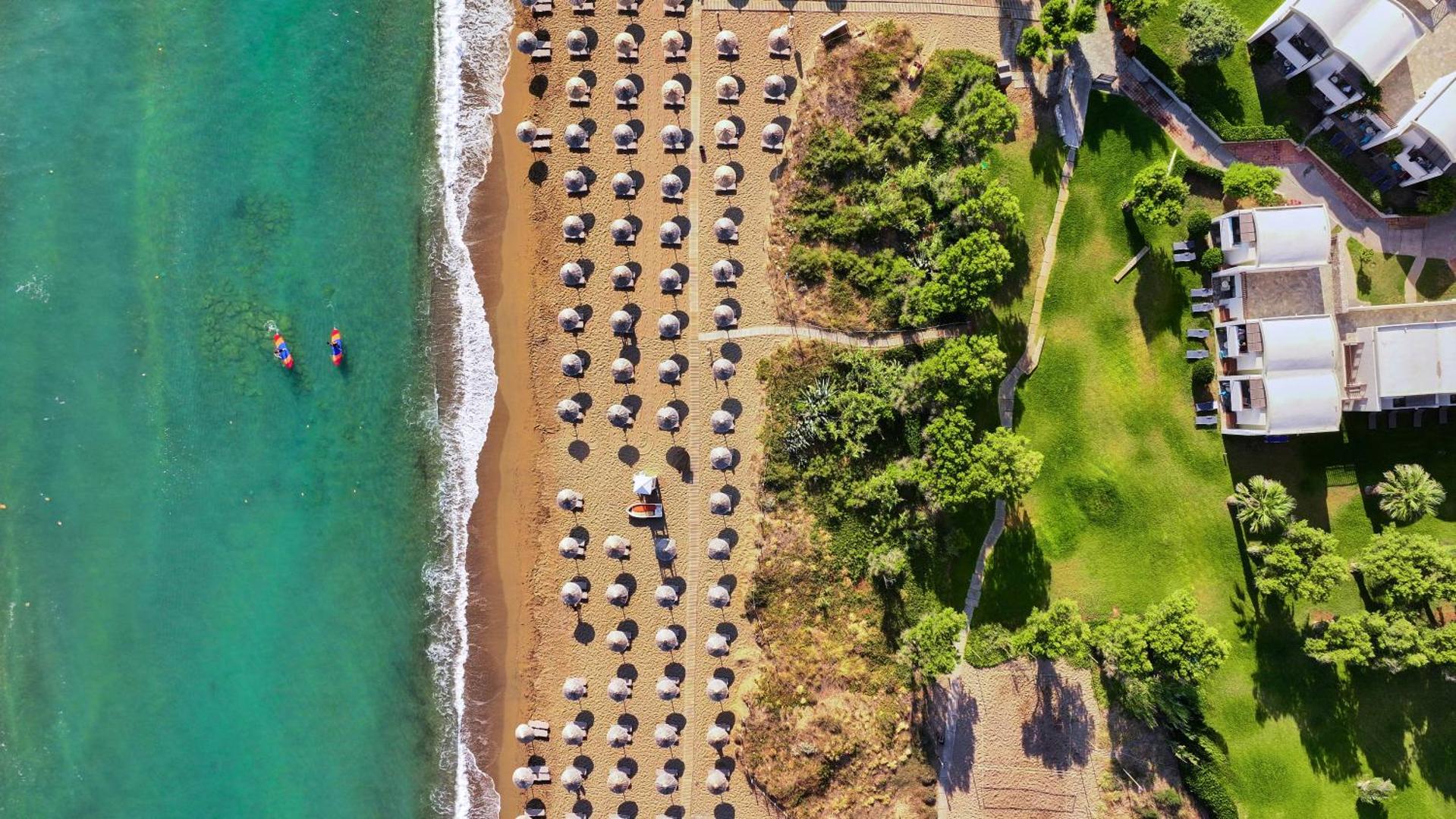 Agapi Beach Resort skats no putna lidojuma uz smilšu pludmali, tirkīza jūru, saulessargiem un viesnīcas dārzu Krētā