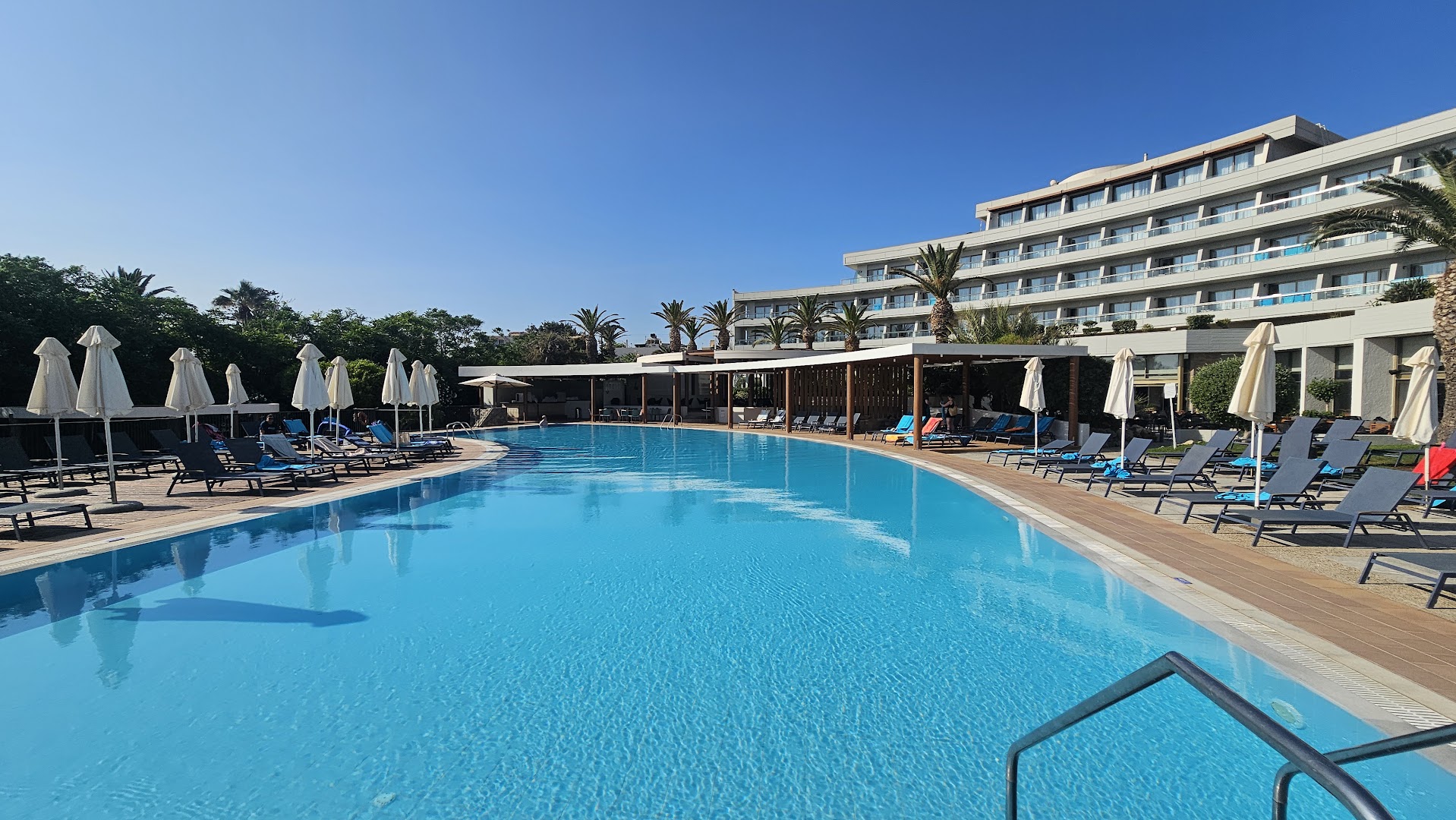 Agapi Beach Resort lielais āra baseins ar skatu uz viesnīcas galveno ēku, palmām un daudziem sauļošanās krēsliem