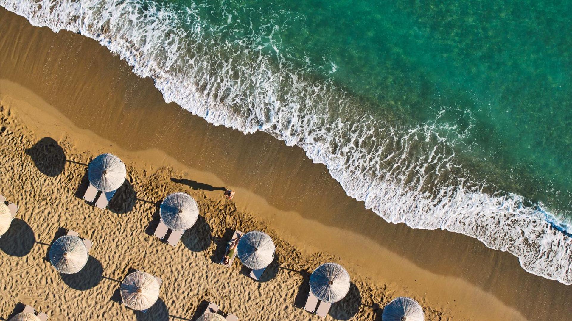 Tuvplāns no augšas uz Agapi Beach Resort pludmales zonu ar saulessargiem un jūras viļņiem smiltīs