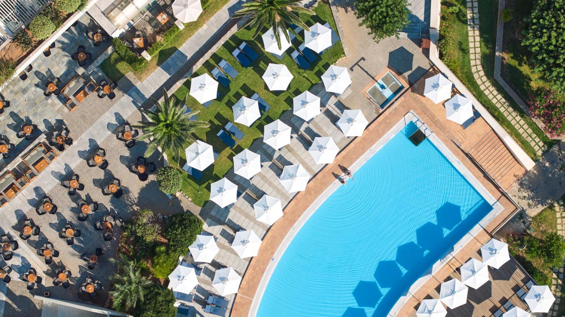 Agapi Beach Resort teritorijas skats no augšas — baseins, restorāna āra terase ar galdiņiem un dārzs ar palmām